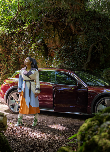 The Founder and Creative Director Priya Ahluwalia stands in a forest in front of the Taycan Cross Turismo.