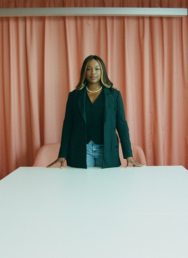 Sharmadean Reid stands at the head of a conference table in front of a soft pink backdrop.