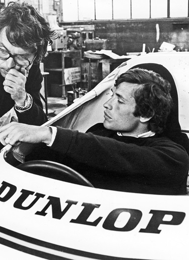 Jacky Ickx sits in a racing car and points to something outside the car which can’t be seen in the picture.