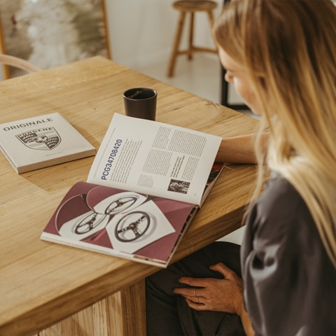Blonde woman reading open Porsche magazine on wooden table