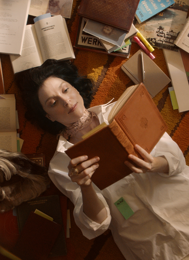 St. Vincent lies on a brightly-carpeted floor in a cozy space. She is surrounded by books and reading a novel.