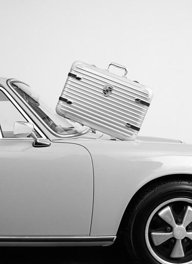 Metal RIMOWA suitcase with Porsche badging leaning on 911